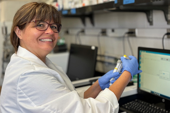 A woman with glasses smiling while holding an object in her gloved hands