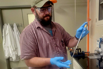 A bearded man wearing glasses and blue nitrile gloves displaying a long object in a lab