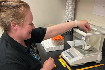 A smiling woman in a black shirt extending her arm into a lab equipment