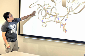 A man in glasses and a grey t-shirt points out a chemical illustration in a Powerpoint presentation